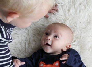Baby lying on sheepskin rug, wearing jumper with fox on it, looking up in adoration at his older brother