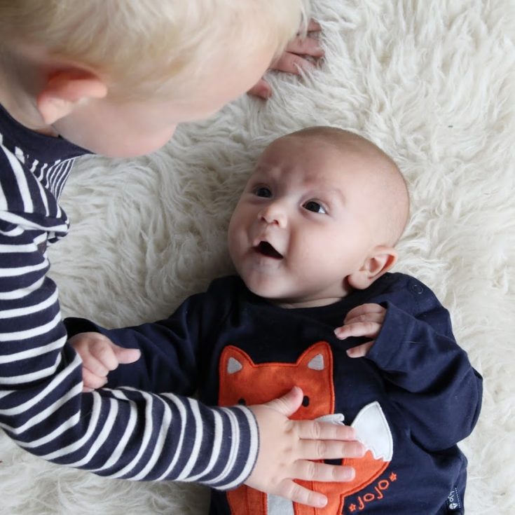 Baby lying on sheepskin rug, wearing jumper with fox on it, looking up in adoration at his older brother