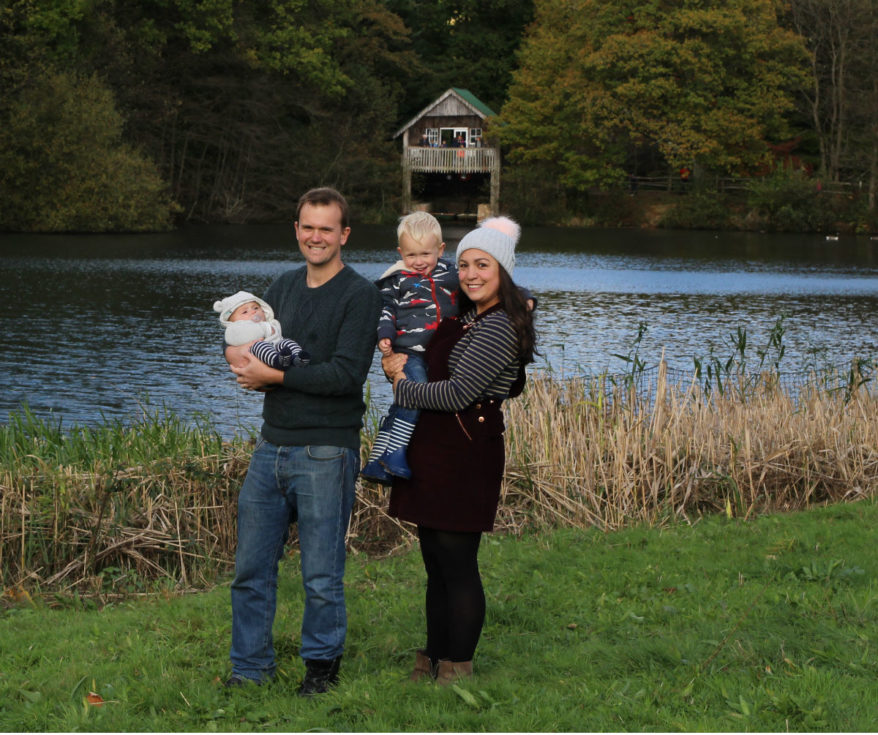 John holding Ben, Katy holding Max, stood in front of lake with boathouse and green and yellow trees in the background