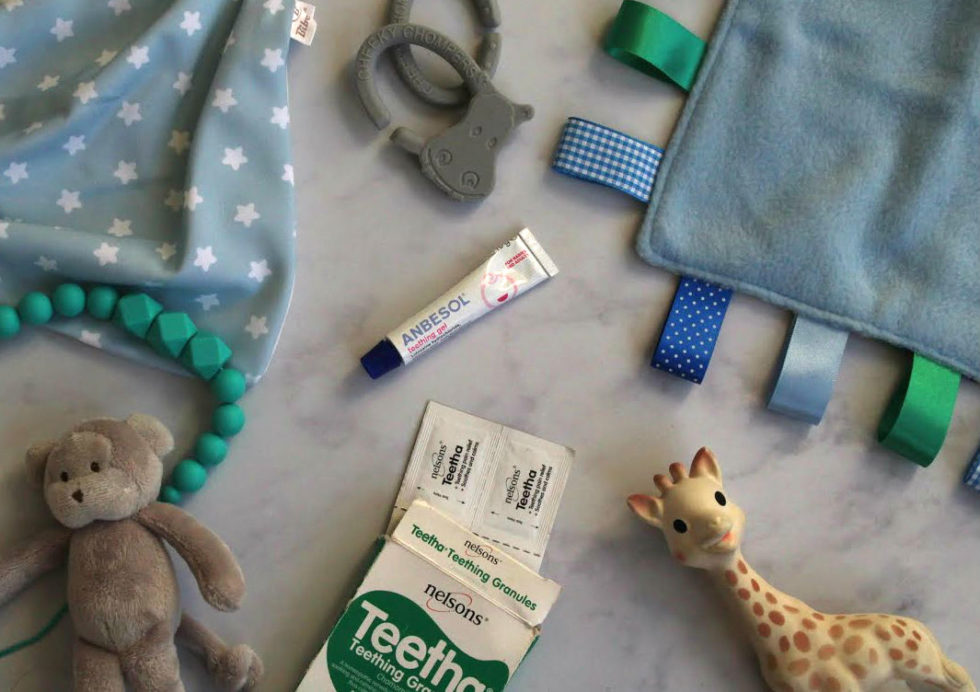 Flat lay of teething products set out on marble worktop: Teetha teething granules, Sophie la Giraffe, Bibetta dribble bib, Anbesol, with taggie blanket and teddy bear