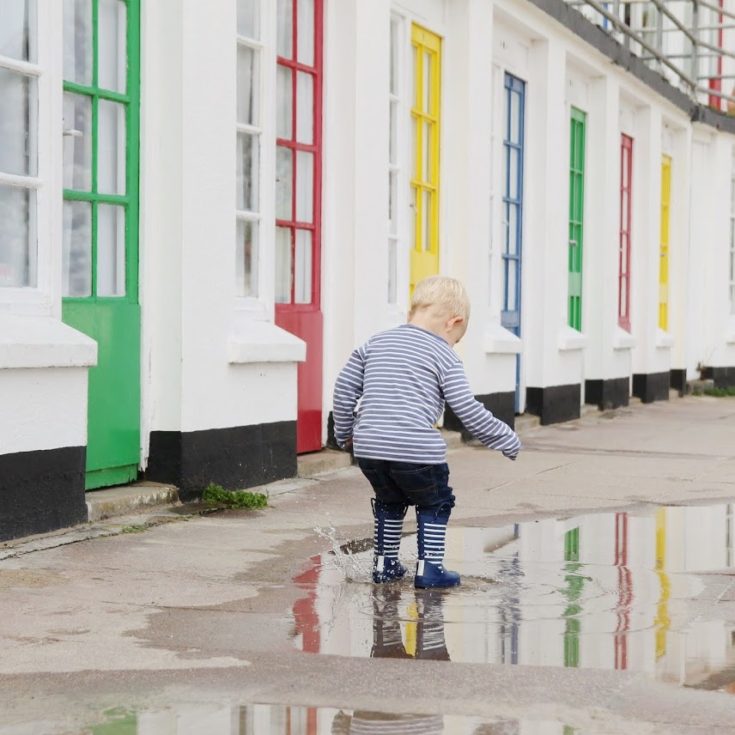 Max jumping in rain puddle, wearing blue stipey wellies, in front of colourful beach hut doors which you can see in the reflection
