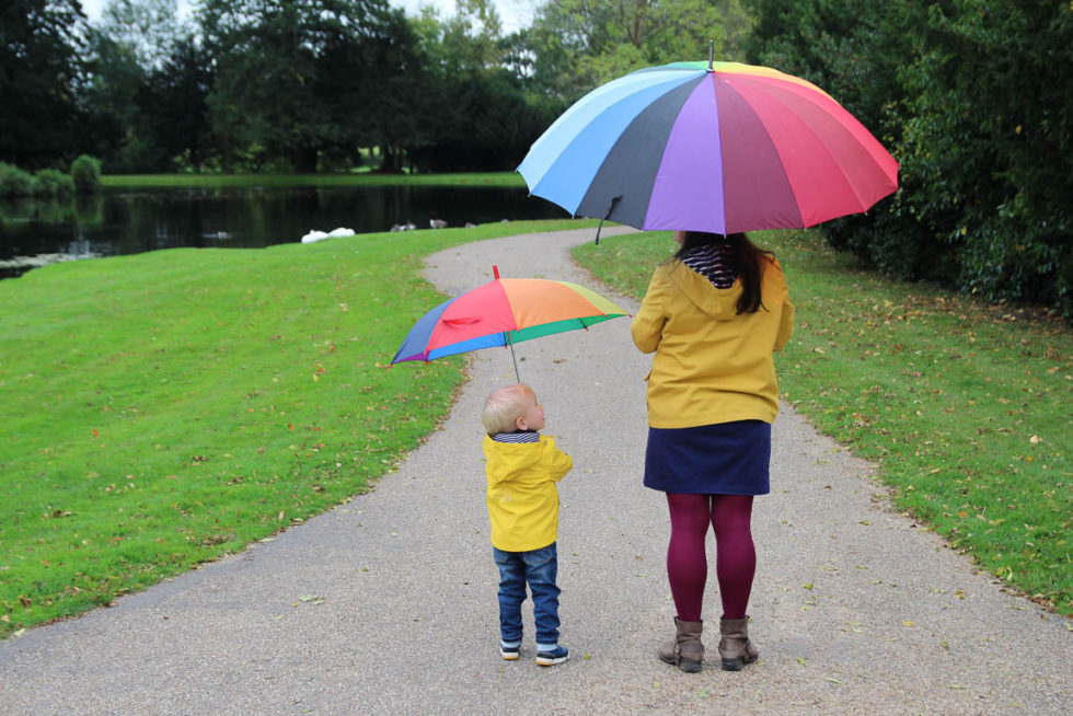 Woman and little boy standing with backs to camera, wearing yellow raincoats and holding yellow umbrellas. The little boy is looking up at his mum