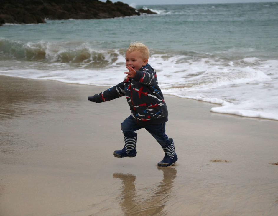 Little boy on beach, dressed in wellies and navy fleece top, running away from the waves at the edge of the sea