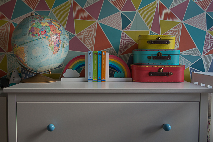 Chest of drawers with blue ceramic knobs, with antique globe, rainbow bookends, and stack of three coloured suitcases on top, with geometric rainbow coloured wallpaper behind