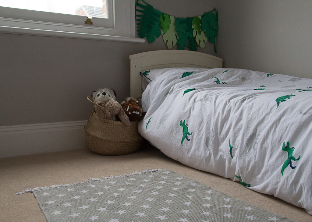 Little boy's bed, with white duvet with dinosaur print, grey bedroom walls, grey star print rug on floor, and basket full of cuddly toys on the floor