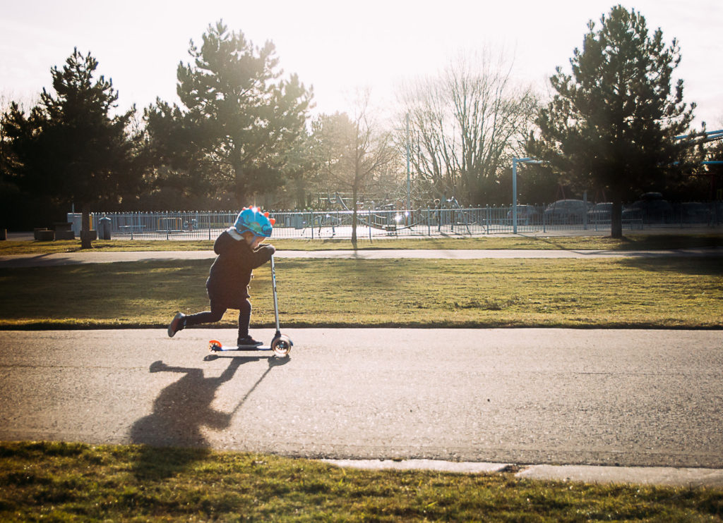 Little boy on scooter with leg in the air, long shadow on the ground in front of him and late evening sun behind him