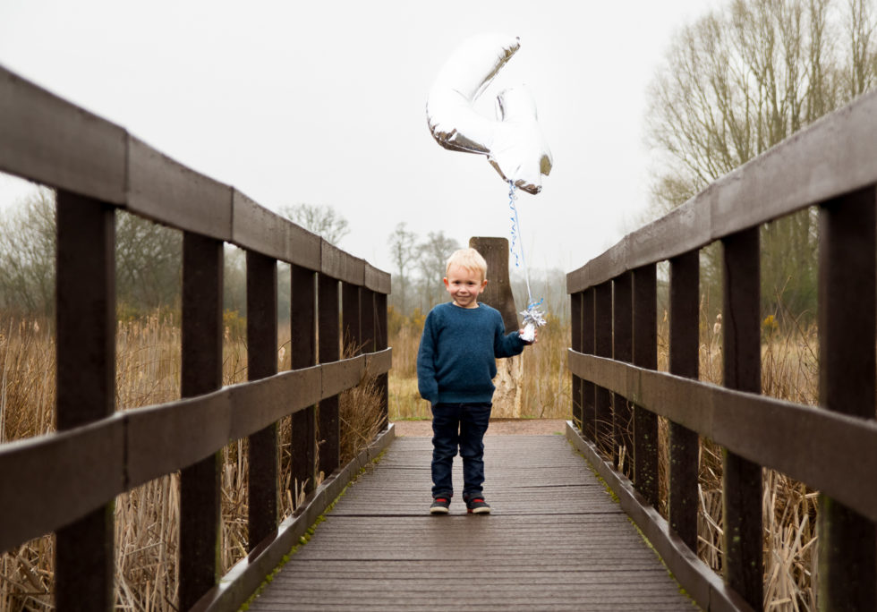 A letter to my son on his 4th birthday. Little boy dressed in green knitted jumper, standing on wooden bridge holding a big 4 silver balloon