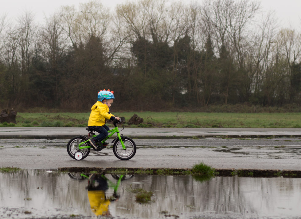 Little boy dressed in yellow raincoat and blue cycle helmet, riding green Frog bike on bicycle track. There is a big puddle and you can see his reflection in it