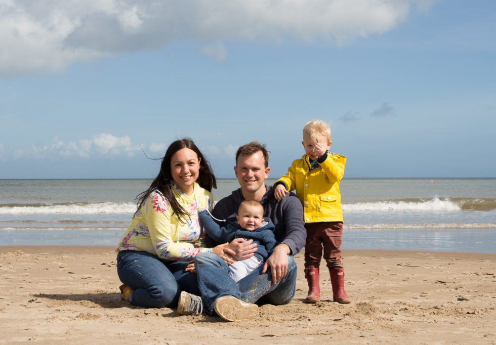 Family sat on the beach. Katy dressed in yellow floral hoodie and jeans, Max in yellow raincoat, John holding baby Ben