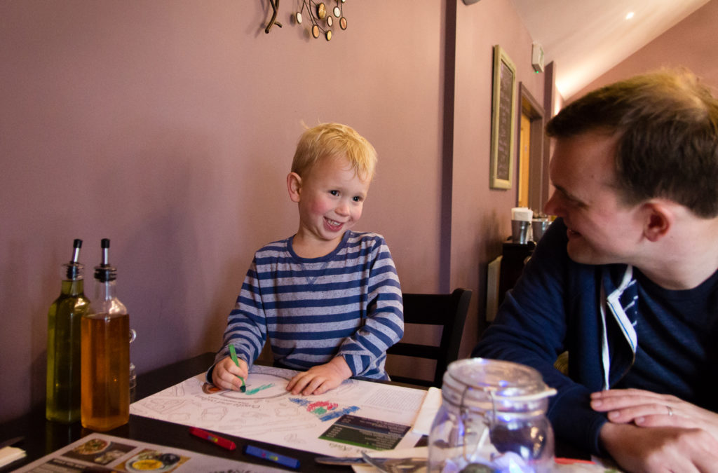 Eating out at The Oak Tree Restaurant at Bluestone Wales. Little boy colouring in and looking at his Daddy and smiling