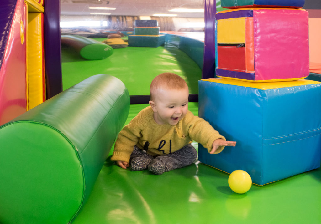 Baby sat in soft play area, with coloured foam stacking bricks next to him. He is reaching for a yellow ball in front of him, smiling