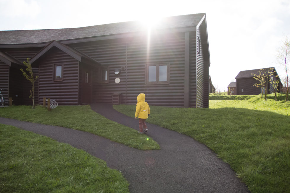 Ramsey lodge at Bluestone Wales. Little boy dressed in yellow raincoat walking up the path towards the front door. The sun is peeking over the roof leaving a sun flare
