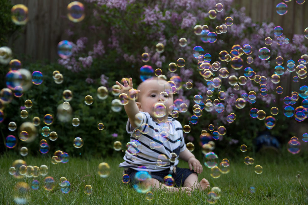Baby sat on grass in front of lilac bush, surrounded by bubbles. He is reaching out with his hand to try to grab a bubble