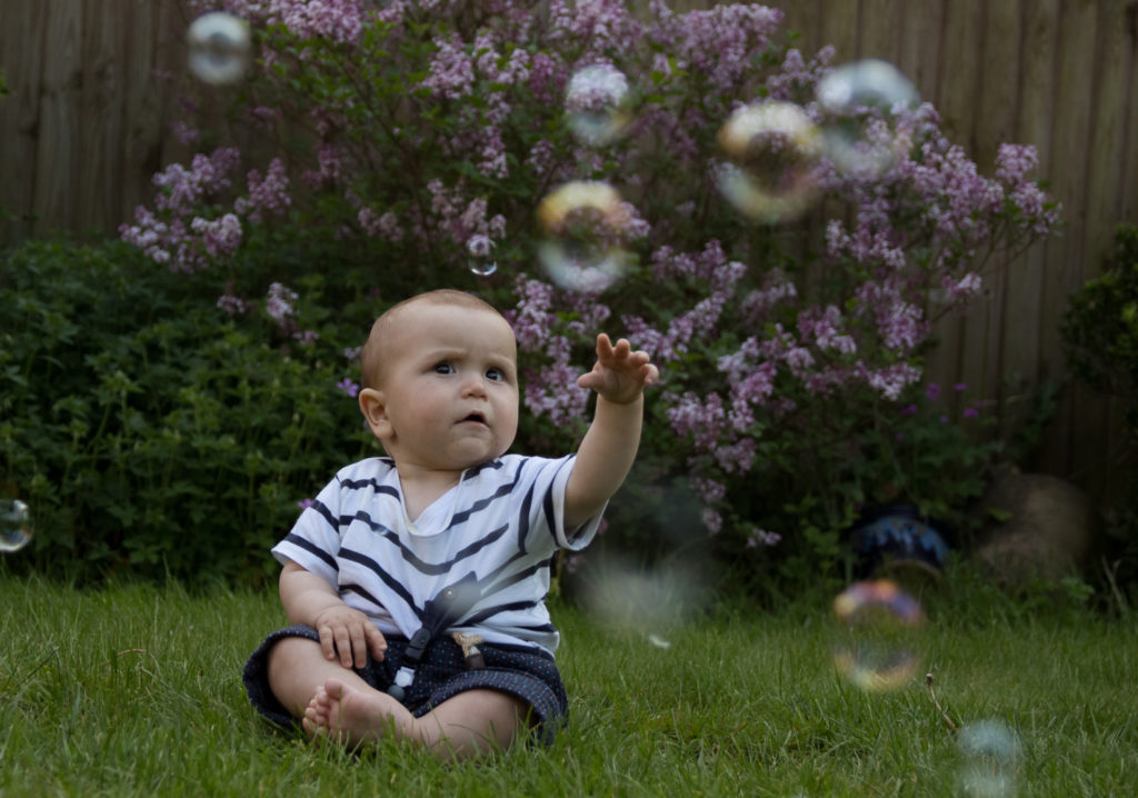 Baby sat on grass in front of lilac bush, with bubbles floating in front of him. He is reaching up with his hand to try to grab a bubble