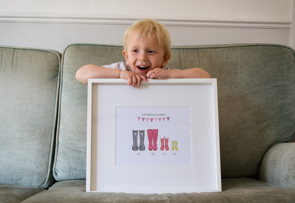 Max sat on the sofa, holding up the framed personalised wellington boot print from Created by Magic