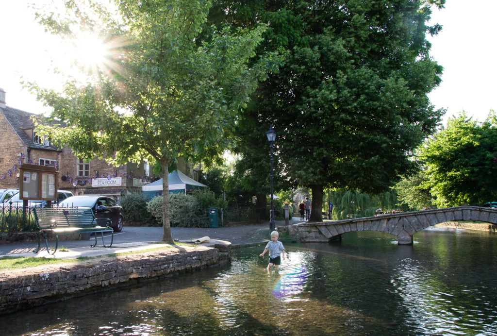 Little boy paddling in the river at Bourton on the Water, with the sun bursting through the trees