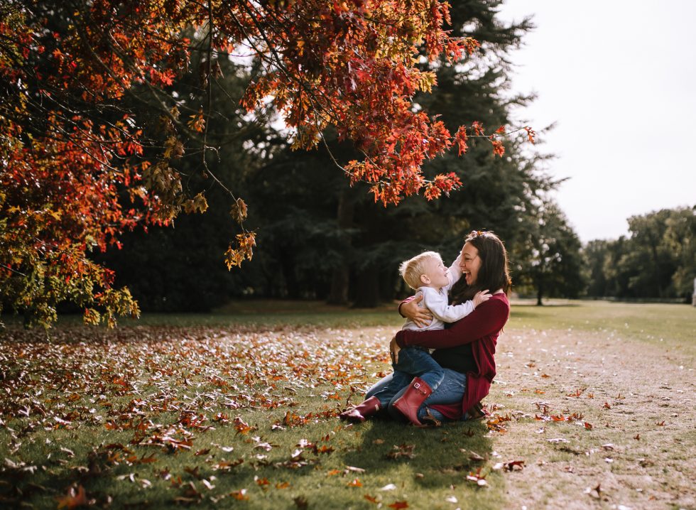 Mother and little boy hugging underneath a tree in autumn sunshine. The leaves on the tree are red, and both mother and son are laughing