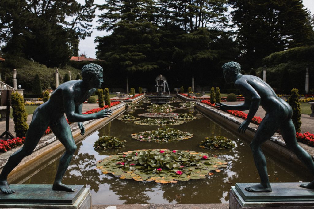 View of the Italian Garden at Compton Acres. A long rectangular pond stretches out ahead, and in the foreground there are two statues of men, facing each other, as if they're ready to fight. The pond is filled with water lillies and lined with pretty red flowers.
