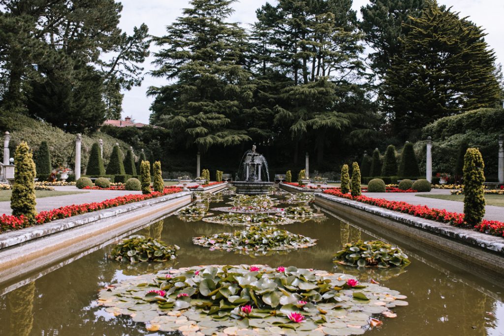 View of the Italian Garden at Compton Acres. A long rectangular pond stretches out ahead, filled with water lillies and lined with pretty red flowers. At the far end of the pond you can see a tall fountain