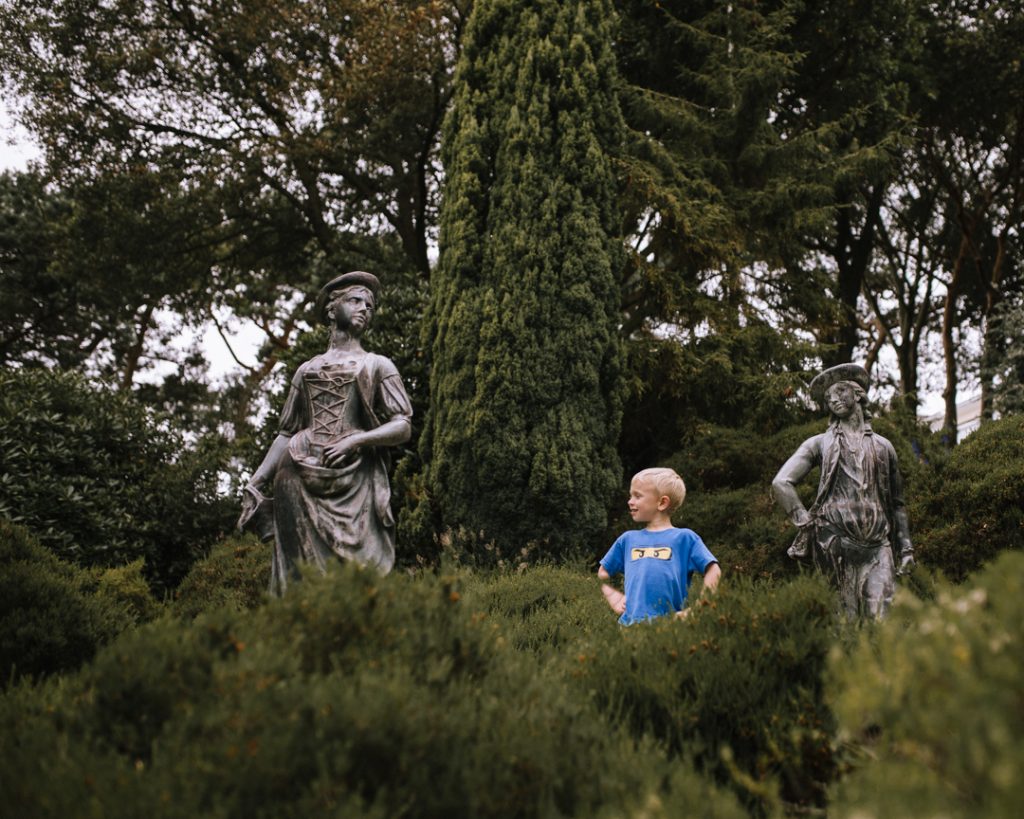 Two grey statues of a man and woman in 18th Century dress, with little boy stood in between them, looking up at them
