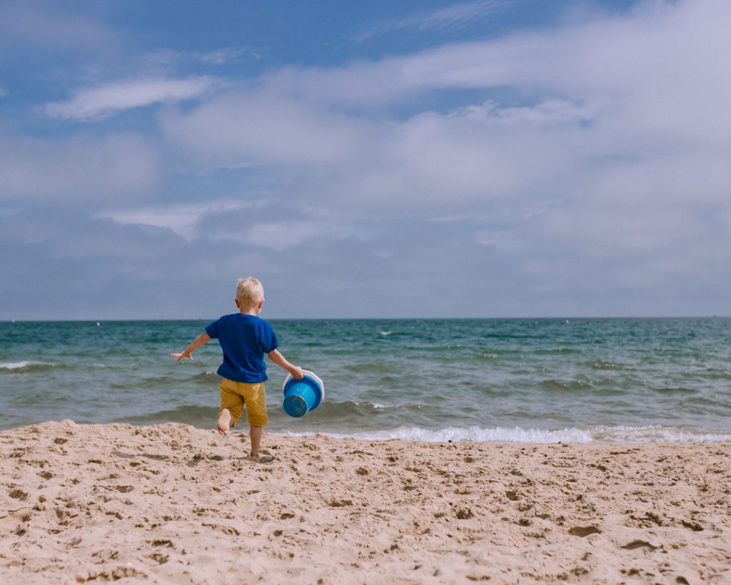 View of the sea, with sandy beach in the foreground and turquoise sea behind. A little boy is running towards the sea, holding a blue bucket