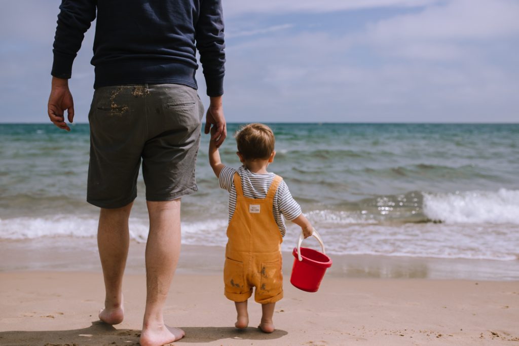 Father and toddler son, holding hands and walking towards the sea. The little boy is wearing yellow dungarees and holding a red bucket