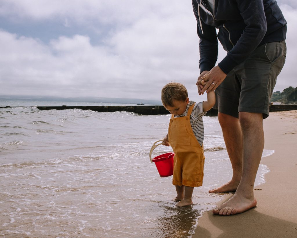 The edge of the sea at Alum Chine beach. A toddler is padding in the sea, holding a red bucket and wearing yellow dungarees. His hand is held by his dad