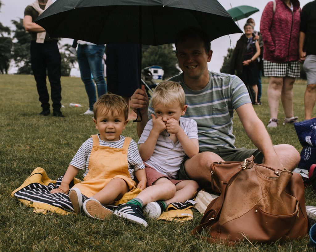 Dad and two boys sat on coats on the grass, with umbrella held over their heads